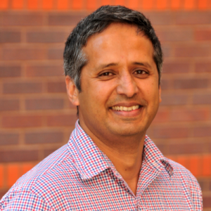 A man in checkered a button up shirt stands and smiles in front of an orange brick wall.