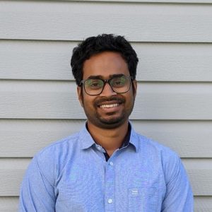 A man with glasses wearing a light blue button up smiles.