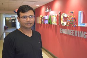 A man in glasses wearing a black shirt stands in front of a mechanical engineering sign.