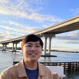 A man in a blue shirt and beige jacket smiles for photo in front of large bridge over water behind him.