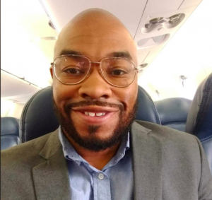 A man in a grey suit and blue shirt wearing glasses smiles while sitting in an airplane seat.
