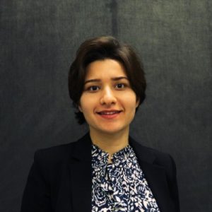 Mojdeh smiles for photo with black, blue, and white shirt under a black sweater. She stands in front of a black wall.