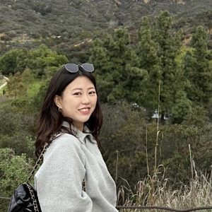Bitgoeul Kim smiles for camera in a grey sweater and sunglasses on her head. She is standing on a hillside with green trees filling the background.