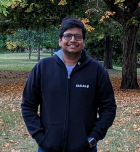 Davesh Lohia smiles for photo wearing glasses in black sweater. Fall leaves and trees are in the background.