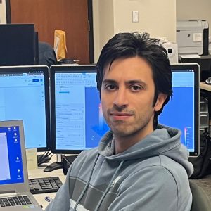 Mehdi Shadkhah looks at camera while sitting in front of laptop and 2 computer screens.