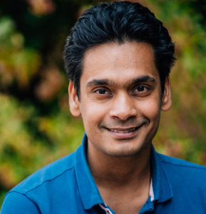 Ronak Tali smiles for camera with blue Polo shirt and green shrubs in background.