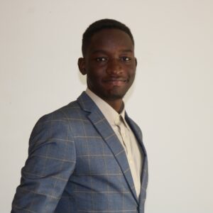 Elvis Kimara stands in front of a white wall and smiles at the camera.