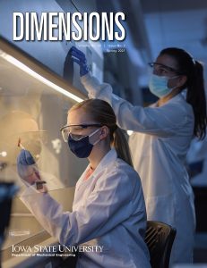 A cover for the Spring 2021 edition of Dimensions, the alumni magazine for mechanical engineering at Iowa State University. In this photo two female students are wearing lab coats, safety glasses, face masks, and rubber gloves. One students examines a 3D-printed sensor under a hood while the other students adjust settings for the hood.