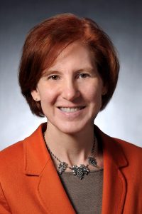 A headshot of mechanical engineering department chair Caroline Hayes. She is smiling and wearing an orange jacket with a brown undershirt.