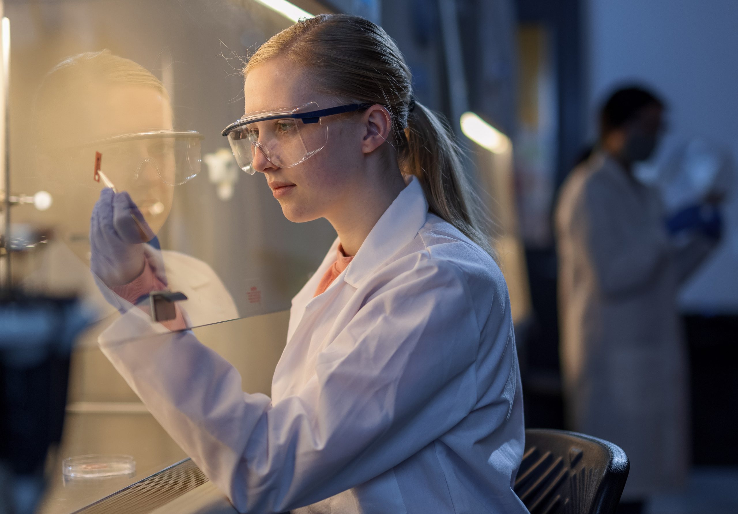 A young lady examines a printed microchip underneath a hood as part of a research experiment. Another female researcher can be seen in the background of the shot.