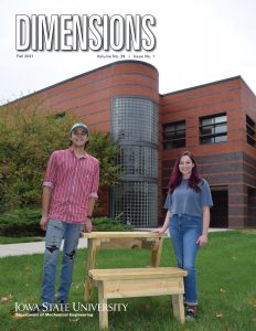 Two college students stand next to a wooden desk they helped to fabricate. The students are standing outside of Black Engineering Building .